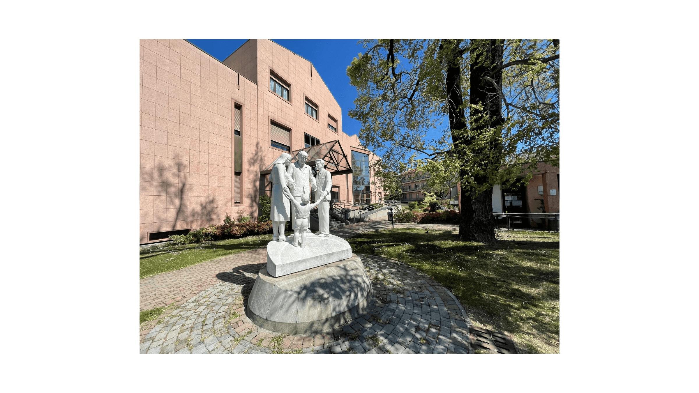 Edificio ospedaliero circondato da un’area verde, con una scultura in primo piano raffigurante operatori sanitari e un bambino.